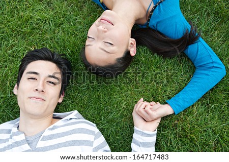 Over head close up portrait view of a young and attractive Japanese couple laying down on green grass and holding hands while relaxing together outdoors.