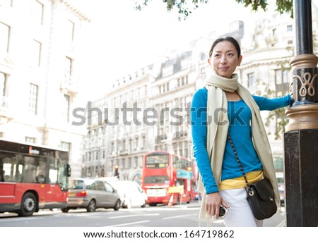 Attractive Japanese tourist woman enjoying sightseeing in a classic architecture street in the destination city of London, carrying a digital photo camera during a sunny day on vacation, outdoors.