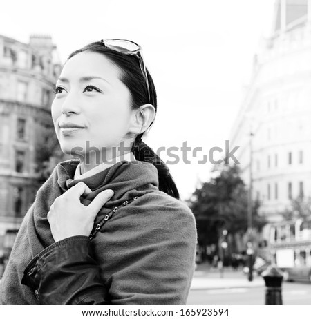 Black and white portrait of a smart and attractive Japanese tourist woman enjoying a visit to a London city street, contemplating and sightseeing during a sunny day on vacation, smiling outdoors.