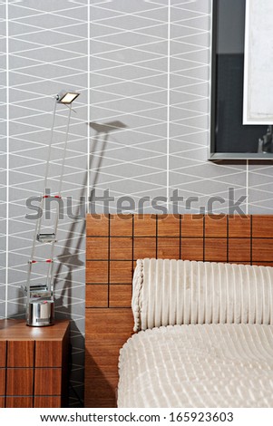 Still life view of a stylish and luxury bedroom with a wooden bed frame and a picture on the wall, showing graphic masculine wallpaper and a bedside table with lamp. Interior with no people.