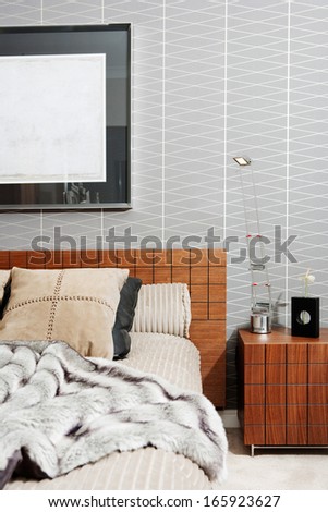 Still life view of a stylish and luxury bedroom with a wooden bed frame and a picture on the wall, showing graphic masculine wallpaper and a fake fur blanket. Interior with no people.