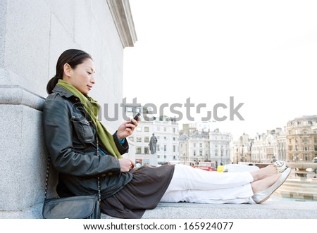 Side view of an attractive Japanese tourist woman relaxing in Traffalgar Square landmark in the city of London, holding a smart mobile phone and relaxing during a sunny day on holiday, smiling.