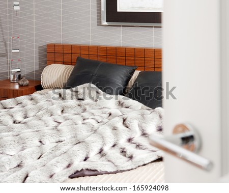Still life view through an open door of a stylish and luxury bedroom with a wooden bed frame and a picture on the wall, showing graphic wallpaper and a fake fur blanket. Interior with no people.