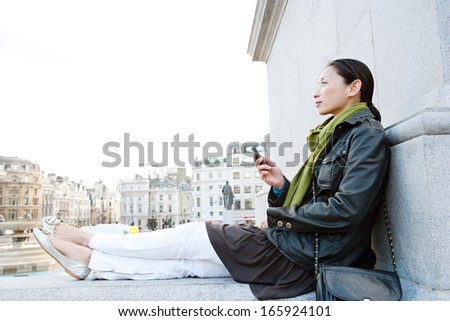 Side view of an attractive Japanese tourist woman sitting and relaxing in Traffalgar Square landmark in the city of London, holding a smart mobile phone and relaxing on vacation.