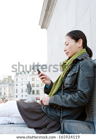 Side view of an attractive Japanese tourist woman relaxing in Traffalgar Square landmark in the city of London, holding a smart mobile phone and relaxing during a sunny day on holiday, smiling.