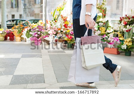 Side view of the lower body section of a young woman walking passed a fresh flowers market stall carrying paper shopping bags during a sunny day outdoors. Faceless figure.
