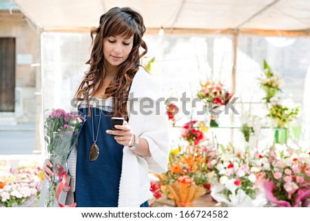 Young consumer woman buying and holding a bouquet of fresh flowers from a floral market stall business and using a smartphone in the store during a sunny day, outdoors.