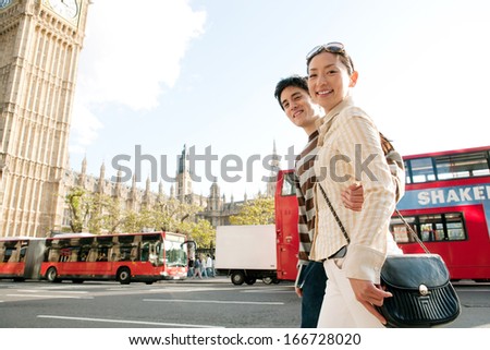 Side view of an attractive and joyful Japanese tourist couple walking passed the Big Ben parliament building in the city of London while on vacation during a sunny day, outdoors.