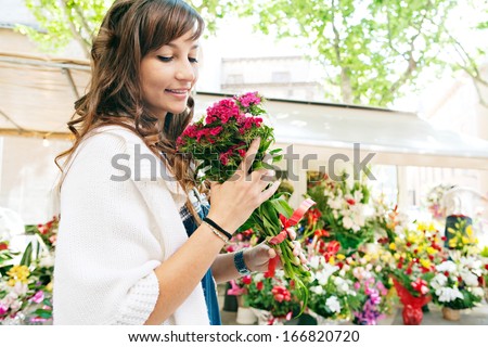 Close up profile portrait of a beautiful and young woman enjoying and smelling a bouquet of flowers while standing in a fresh floral market stall during a sunny day outdoors.