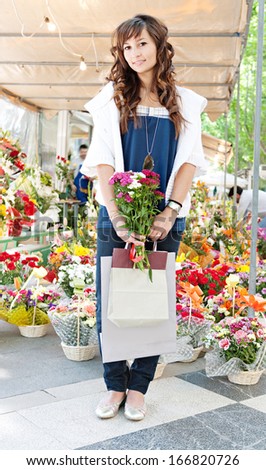 Attractive young woman shopping in an outdoors flowers market stall, buying a small bouquet of flowers from the colorful selection of arrangements and carrying paper shopping bags.