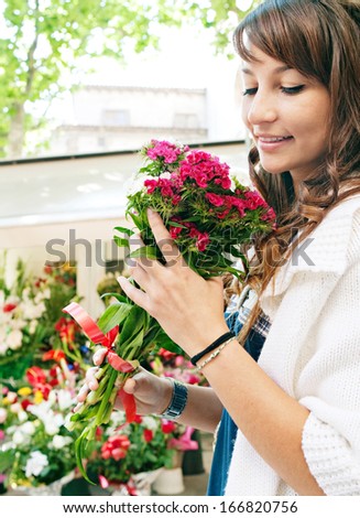Close up profile portrait of a beautiful and young woman enjoying and smelling a bouquet of flowers while standing in a fresh floral market stall during a sunny day outdoors.