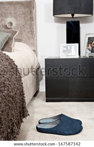 Still life view of an elegant master bed in a design home bedroom with warm colors and fabrics, and a pair of blue slippers resting on the beige carpet floor. Home interior view with no people.