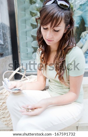 Portrait of a fashionable young woman sitting by a fashion store window display counting bank notes bills and coins currency money during a shopping day out, outdoors.