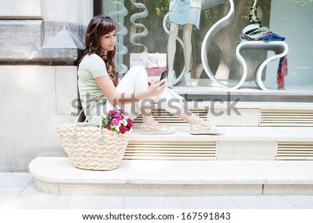 Side view of an attractive young woman sitting on the steps of a fashion store window display with manikins, holding and using her smartphone during a shopping day out, outdoors.