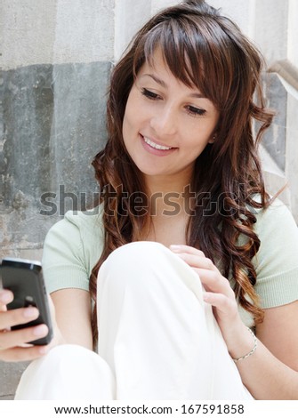 Close up portrait of an attractive teenager woman networking and using her smartphone while sitting against a stone textured wall during a sunny and fun day out, outdoors.