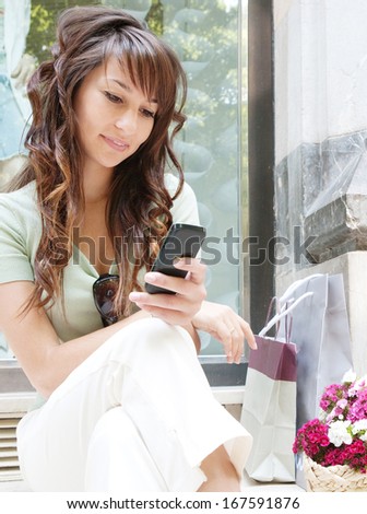 Portrait of a young woman sitting by a fashion store window display relaxing and browsing the internet using her smartphone during a shopping day out, smiling outdoors.