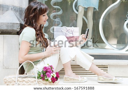 Side view of an attractive young woman sitting on the steps of a fashion store window display with manikins, holding and using her smartphone during a shopping day out, outdoors.