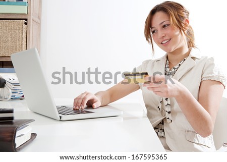 Side portrait view of a young professional business woman using a laptop computer and a credit card to make a payment on line while sitting at her work desk against a white wall. Office interior.