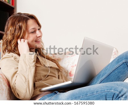 Side portrait of a teenager girl sitting on an armchair in a home living room and using a laptop computer to listen to music with her earphones, smiling and enjoying technology, interior.