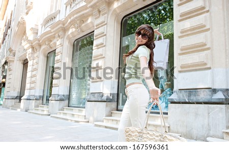Joyful attractive young consumer woman near a shopping mall entrance with elegant buildings and stores, carrying paper bags and dancing having fun to turn at the camera smiling.