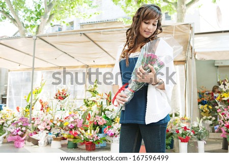 Attractive young woman shopping in an outdoors fresh flowers market stall, buying a small bouquet of flowers from the colorful selection of arrangements during a sunny day in the city.