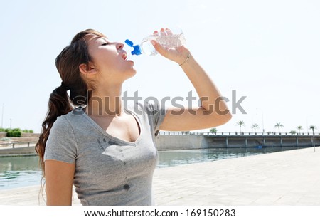 Profile portrait view of a young sporty woman drinking mineral water from a plastic bottle and getting refreshed after exercising outdoors during a sunny summer day.