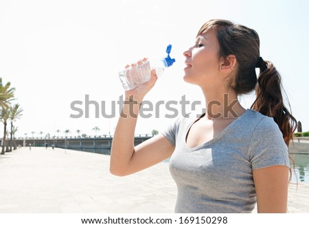 Profile portrait view of a young sporty woman drinking mineral water from a plastic bottle and getting refreshed after exercising outdoors during a sunny summer day.