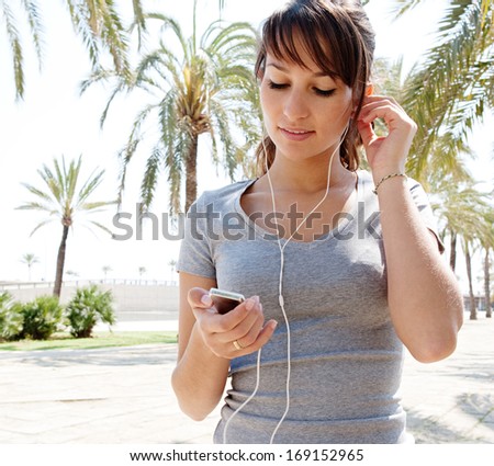 Sporty young woman taking a break from exercising and holding a smartphone and listening to music with her headphones during a sunny day in a park with palm trees and blue sky, outdoors.