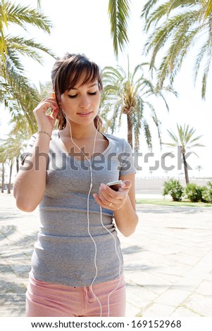 Sporty young woman taking a break from exercising and holding a smartphone and listening to music with her headphones during a sunny day in a park with palm trees and blue sky, outdoors.