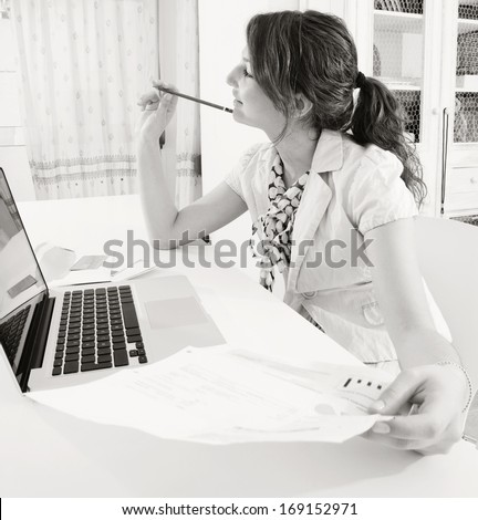 Black and white portrait of a young professional business woman using a laptop computer sitting at her work desk holding utility bills and paying with a credit card on line while being thoughtful.