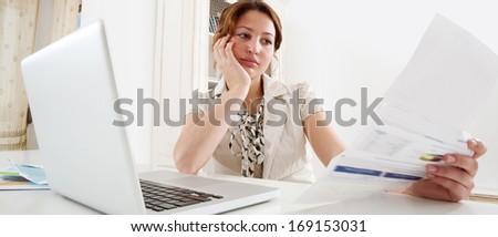 Panoramic Portrait Of A Young Professional Business Woman Using A Laptop Computer Sitting At Her Work Desk Holding Utility Bills And Bank Statements Being Thoughtful And Worried. Office Interior.