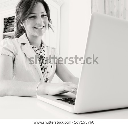 Black and white close up portrait of a young attractive professional business woman sitting at her desk and typing on her laptop computer while smiling and enjoying technology, office interior.
