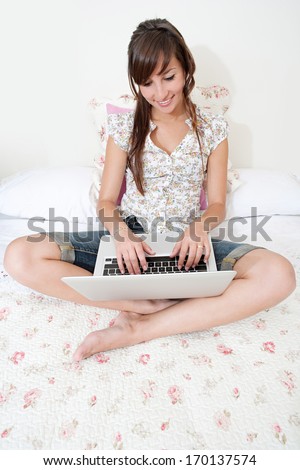 Over head view of an attractive young woman sitting on her bed with crossed legs while using a laptop computer to network with friends. Home interior technology lifestyle.