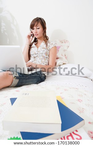 Attractive young student woman woman sitting down and relaxing on her bed at home with books and a laptop computer, browsing on line while having a phone conversation. Home interior.