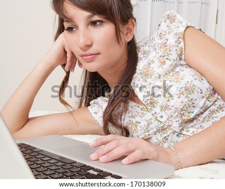 Attractive young woman laying down on her bed at home using a laptop computer and browsing on line. Searching, reading and studying in a home interior.