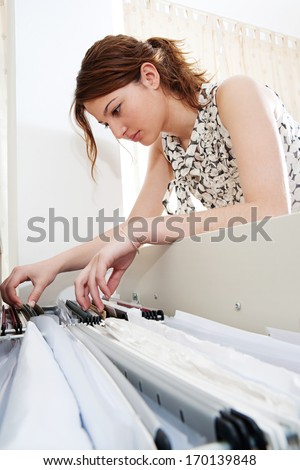 Side portrait view of an attractive office businesswoman flicking through the clients files in an open filing cabinet, searching for and picking a file in an office interior.