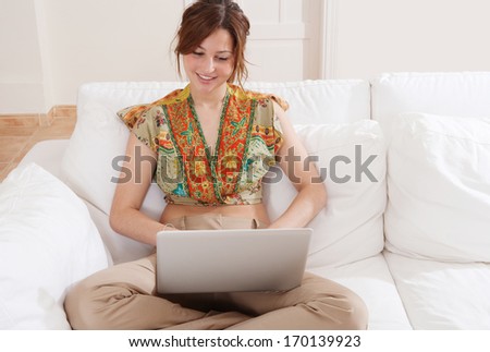 Joyful young woman sitting with crossed legs and relaxing on a home white sofa living room, smiling and using a laptop computer to network on line, sunny home interior.
