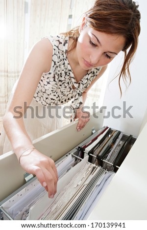 Close up portrait view of an attractive office business woman flicking through her clients files in an open filing cabinet, searching for and picking a file in an office interior during a sunny day.