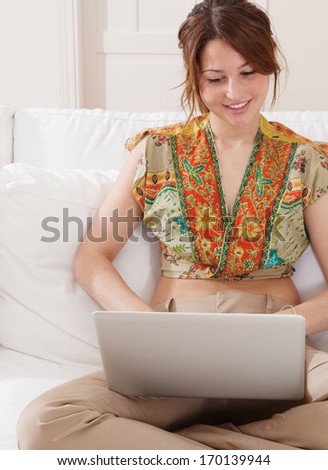 Joyful young woman sitting with crossed legs and relaxing on a home white sofa living room, smiling and using a laptop computer to network on line, sunny home interior.