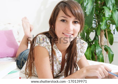 Close up portrait of an attractive student woman laying down on her bed at home while working on her homework from college, preparing for exams and smiling. Home interior.