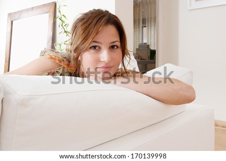 Close up portrait of a beautiful hispanic young woman lounging at home and relaxing on a white sofa during a sunny summer day, being thoughtful and smiling. Home interior.