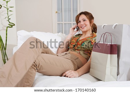 Joyful young woman relaxing on a home white sofa, smiling and having a telephone conversation after a shopping day out with paper bags, home interior.