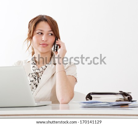 Portrait of a young and attractive professional business woman sitting at her office work desk having a telephone conversation and using a laptop computer, isolated on a white wall. Office interior.