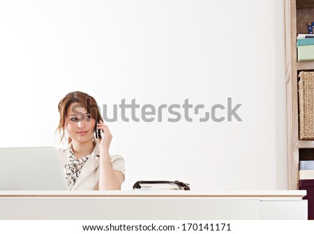 Young and attractive professional businesswoman sitting at her work desk against a white wall and using a laptop computer and a telephone to make a phone call in her office, interior.