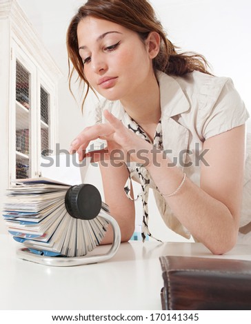 Portrait of a young business woman flicking through her clients contact details database on her roller deck with her fingers while at her working desk, picking a card. Office interior.