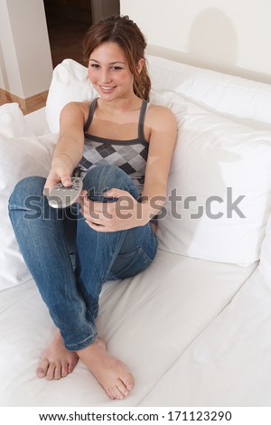 Attractive young woman relaxing and sitting down on a white sofa in a home living room, zapping with a control remote while watching tv at home and smiling. Home technology interior.