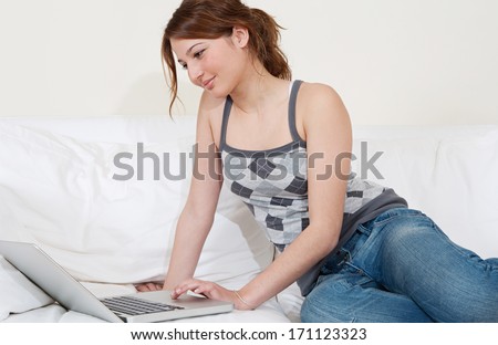 Side view of an attractive young woman lounging and relaxing on a white sofa in a home living room using a laptop computer and smiling. Home technology interior.