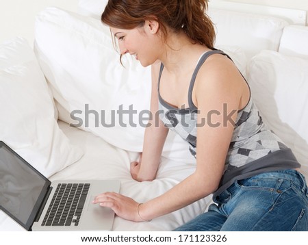 Side view of an attractive young woman lounging and relaxing on a white sofa in a home living room using a laptop computer and smiling. Home technology interior.