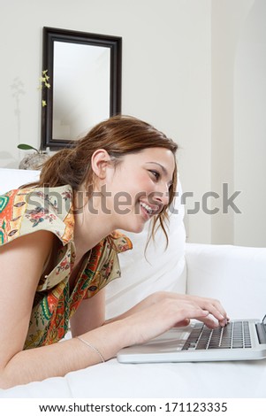 Close up profile portrait view of an attractive young woman using and typing on a laptop computer while laying down on a white sofa at home, relaxing and lounging while networking and smiling.