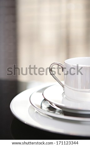 Close up still life detail view of half a serving set of plates and cup for one on a luxury reflective diner table in a quality home with smart curtains. Home interior detail view.